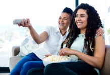 Top 10 LGBTQIA+ Shows & Movies to Watch This Pride Month A happy couple sitting on the couch eating popcorn while searching for a TV show or movie to stream