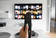 How To Safely Watch 2024’s Most-Anticipated New TV Shows A woman’s hand holding up a TV remote in front of a television screen