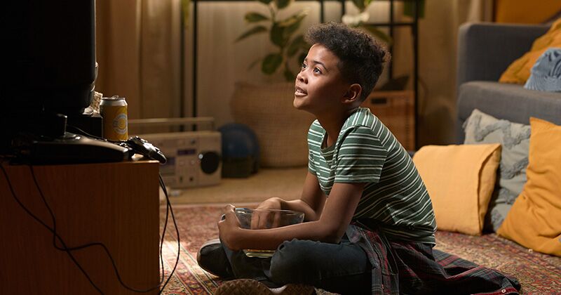 Nostalgia2 African American schoolboy sitting cross-legged on the floor watching television with a popcorn bowl in his lap in a cozy eighties living room