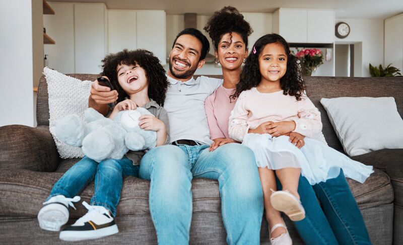 SummerCamp2 Mom, dad and children on sofa, watching tv and happy family bonding together in living room. Smile, happiness and parents relax with kids on couch, streaming television show subscription or movies.
