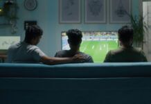 Illegal Sports Streaming Sites to Avoid in 2026 Three young men sitting on the sofa while streaming a soccer match on TV at home.