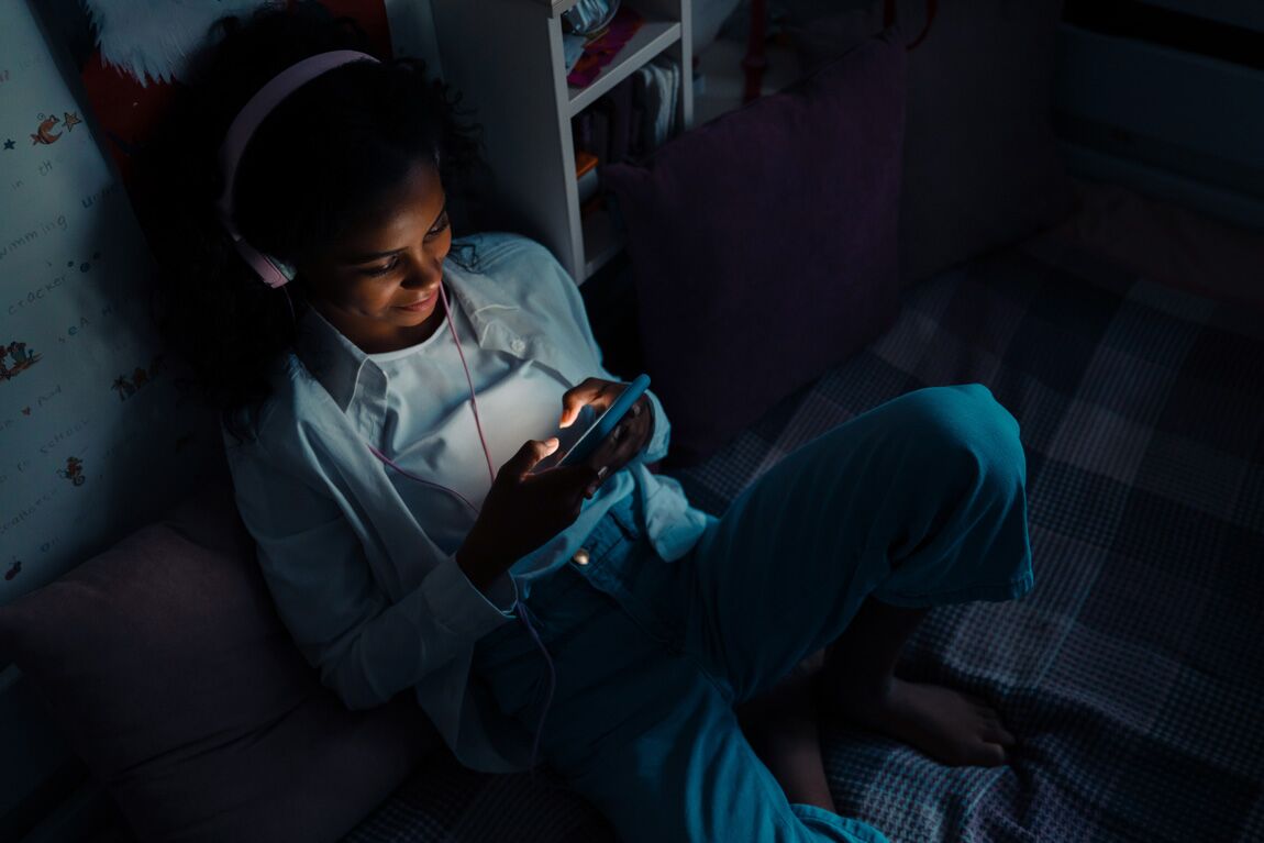 WhyIS_SafeStreaming Smiling teen girl sitting on a bed with phone texting or using mobile apps in her room at home