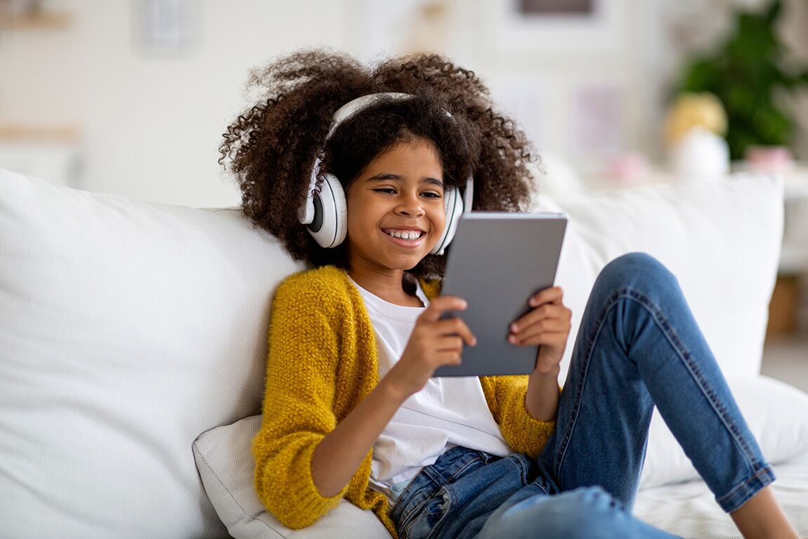 ParentalControl Happy kid school girl sitting on couch, using digital tablet and wireless headset, playing mobile game or checking newest app, home interior, copy space